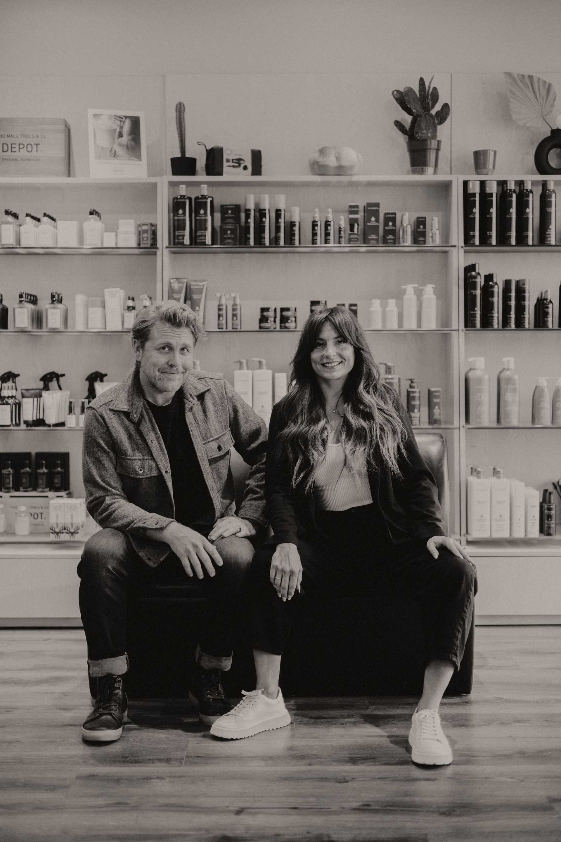 Man and woman smiling, sitting in salon with shelves of hair products behind them.