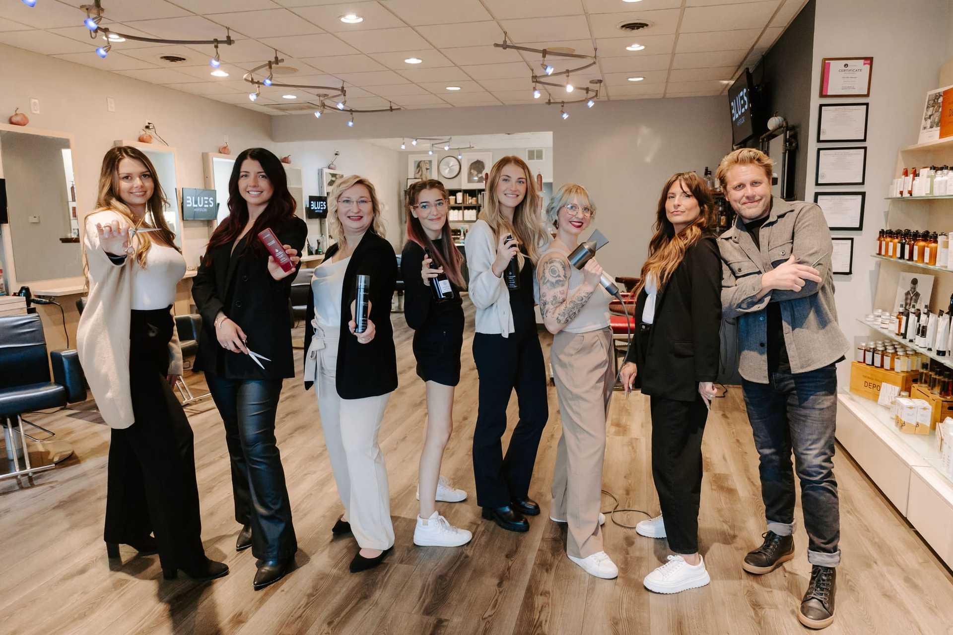 Group of smiling salon staff holding hair styling tools in a modern salon setting.